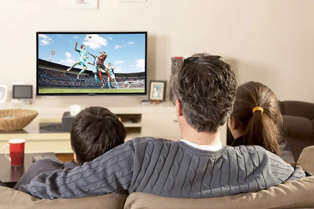 homem e duas crianças sentados em um sofá bege assistindo futebol em frente a uma smart tv 32"