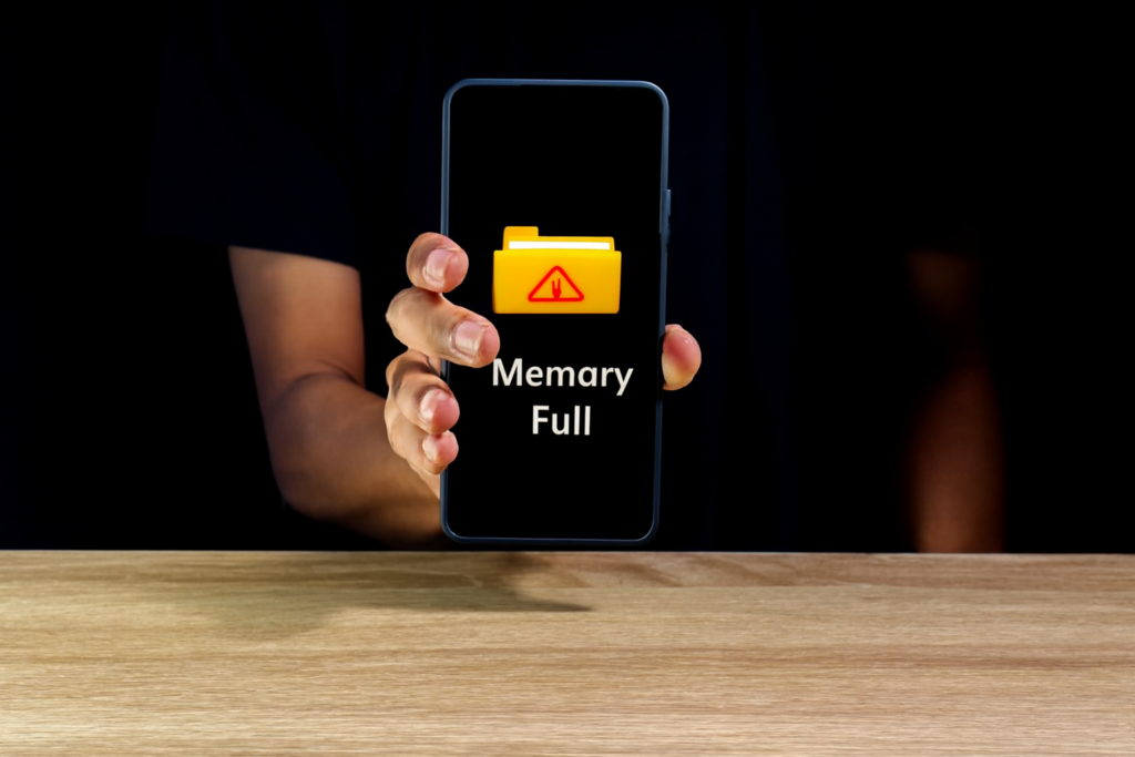 Pessoa segurando um smartphone com a tela exibindo o aviso “Espaço insuficiente”, acompanhado de um ícone de pasta amarela com triângulo de alerta vermelho. Fundo escuro, camisa preta e superfície de madeira em destaque.