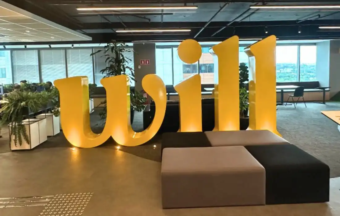 Escultura amarela com a palavra "will" em escritório moderno e iluminado do Will Bank, com móveis modulares e plantas.