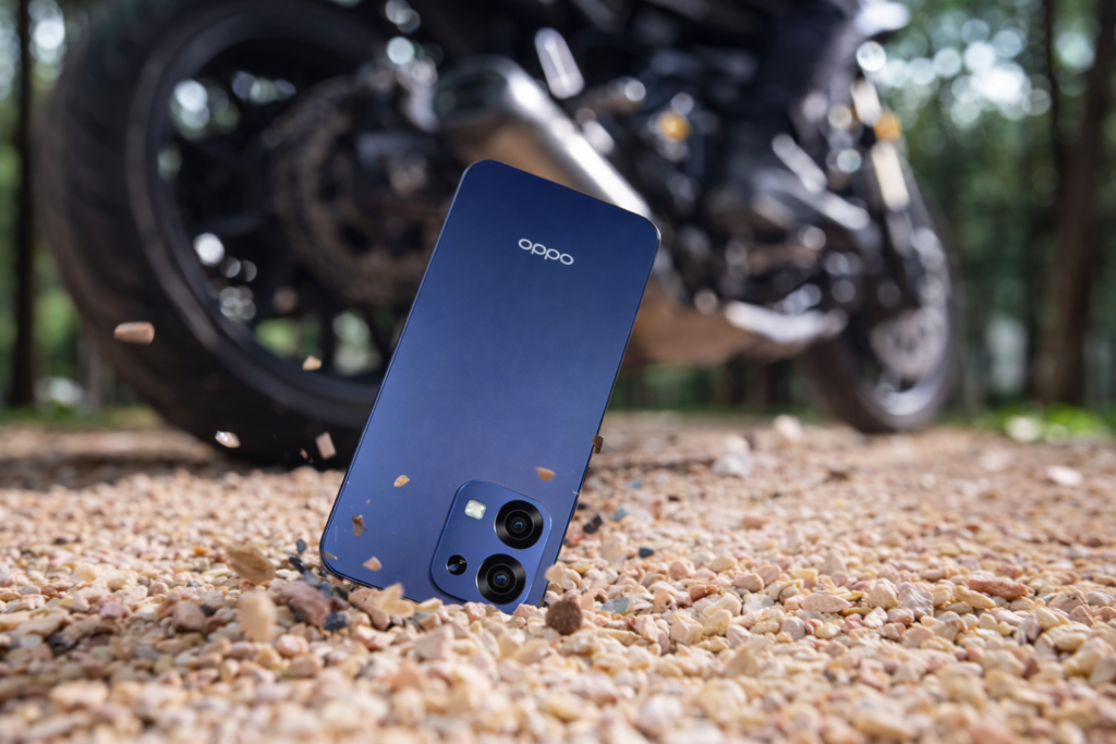 Smartphone Oppo A6 Pro 5G azul espacial em destaque sobre cascalho, com uma motocicleta desfocada ao fundo em ambiente externo.