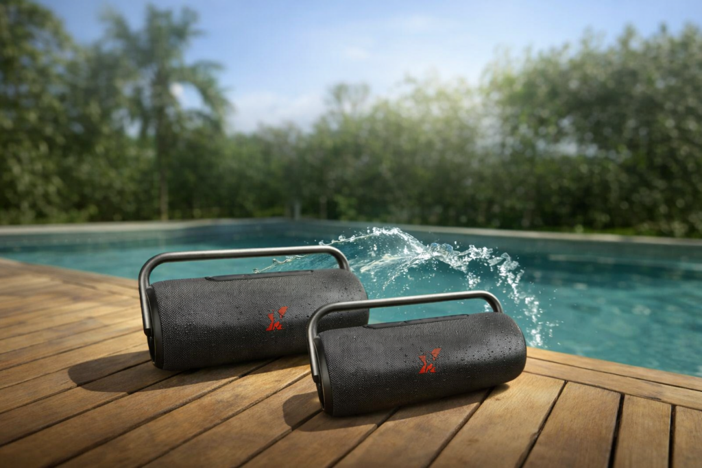 Duas caixas de som port&aacute;teis pretos Amvox ACA 610 Boombox Black sobre deck de madeira &agrave; beira da piscina, com respingos de &aacute;gua e fundo tropical.