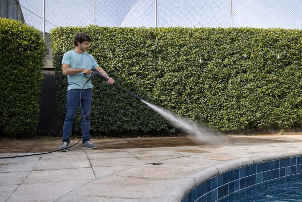 Um homem jovem com cabelo castanho e camiseta verde-&aacute;gua usa uma lavadora de alta press&atilde;o karcher k6 para limpar o piso de pedra ao redor de uma piscina azul. 